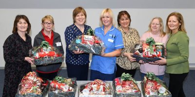 Seven women standing behind a table. They are holding hampers wrapped in festive wrapping. There are more hampers sitting on the table.