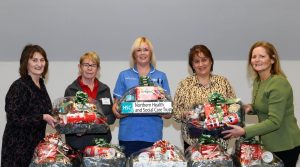 Five women hold festive hampers. They are standing behind a table, with hampers on the table as well.