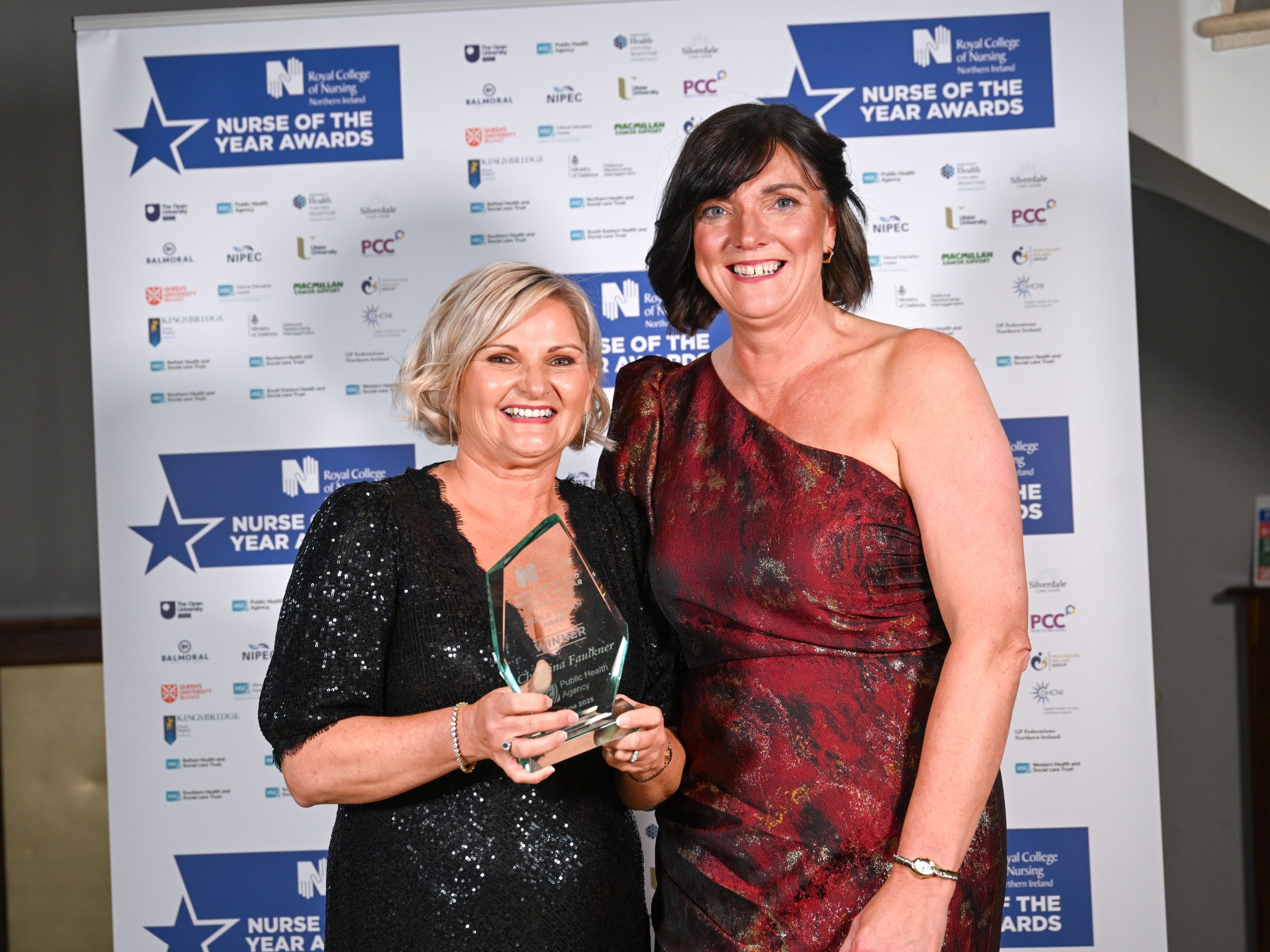 Two women at an awards event, standing in front of a branded backdrop. One is holding a glass trophy.
