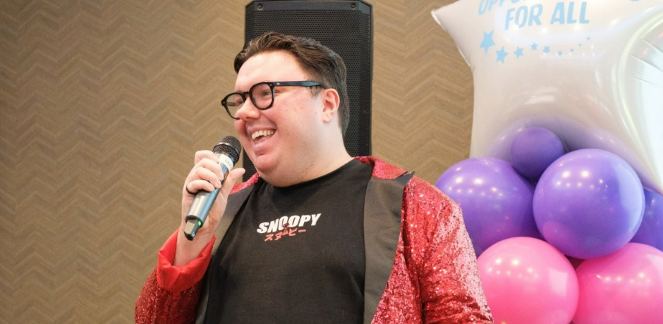 A man wearing a red sparkle jacket holds a microphone. There is a balloon display behind him.