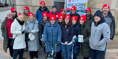 A group of people standing together outside Coleraine Town Hall/ Some are wearing red Northern Ireland Chest, Heart & Stroke branded hats. There is a pop-up banner in the background about Prescribe A Mile.