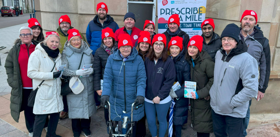 A group of people standing together outside Coleraine Town Hall/ Some are wearing red Northern Ireland Chest, Heart & Stroke branded hats. There is a pop-up banner in the background about Prescribe A Mile.