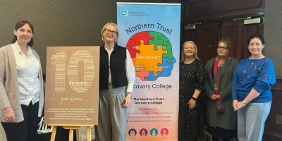 Five women standing together alongside a pop-up banner about the Recovery College and a board featuring a large number 10 and positive words.