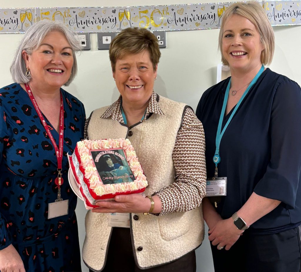 Three women stand beside each other. One is holding a cake.