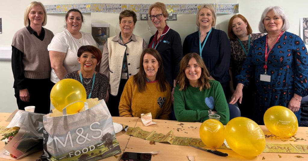 A large group of women, some sitting, some standing, behind a table. There are yellow balloons on the table.