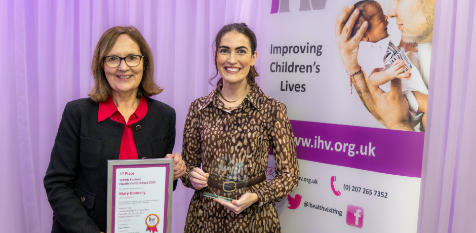 Two women stand beside each other. One is holding a framed certificate and there is a pop-up banner to the side.