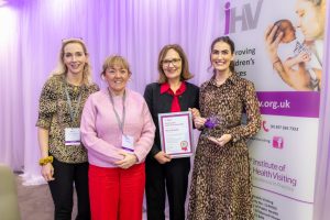 Four women stand beside each other. One is holding a framed certificate and there is a pop-up banner to the side.