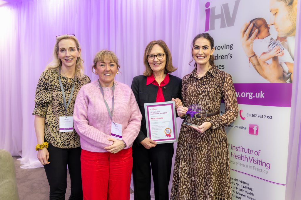 Four women stand beside each other. One is holding a framed certificate and there is a pop-up banner to the side.