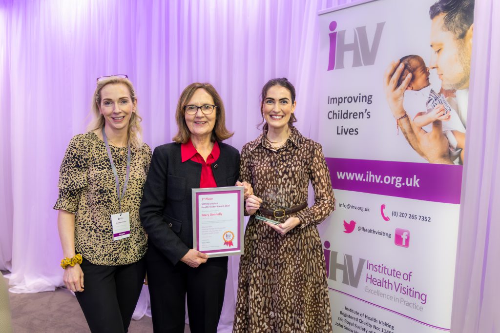 Three women stand beside each other. One is holding a framed certificate and there is a pop-up banner to the side.