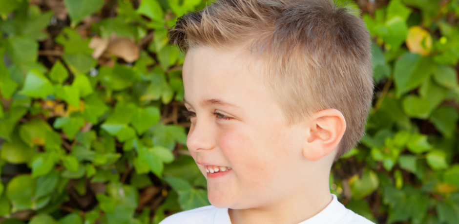 Profile view of a young boy, with a green hedge behind him.