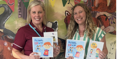 Two women stand beside each other in front of a colourful mural. One woman is wearing a maroon healthcare tunic. They are holding a children's book about FH.
