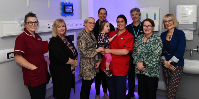 A group of people standing in a clinical setting. A nurse is holding a child in her arms.