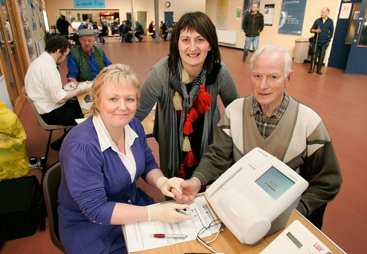 Two women with a man, in a large room. The man is getting a health check.