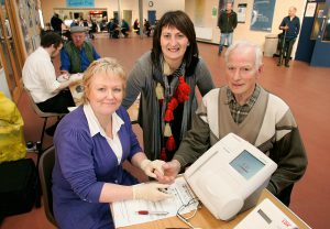 Two women with a man, in a large room. The man is getting a health check.