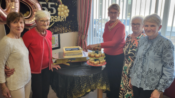 Five women stand beside a table, one is preparing to cut a celebratory cake.