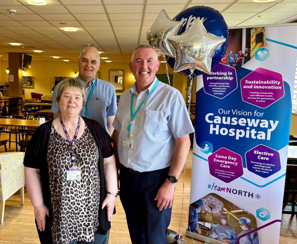 Three people stand smiling in front of a banner detailing the vision for Causeway Hospital, with blue and silver balloons.