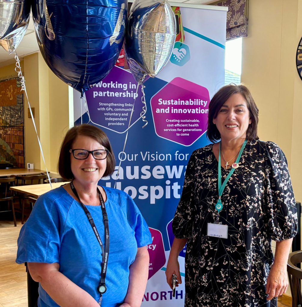 Two women smiling in front of a banner for Causeway Hospital, with balloons in the background.