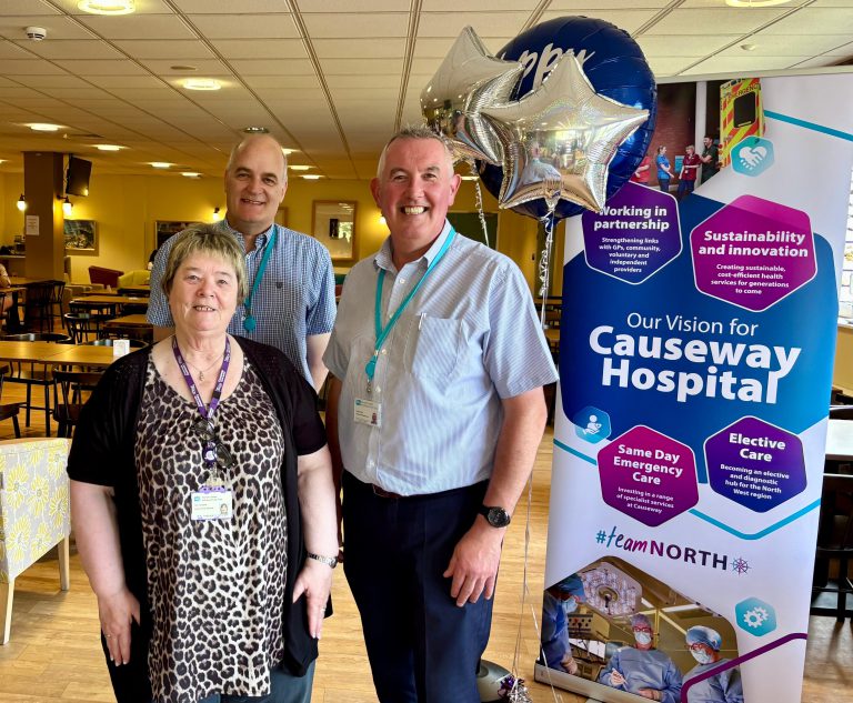 Three people stand smiling in front of a banner detailing the vision for Causeway Hospital, with blue and silver balloons..