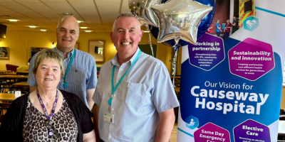 Three people stand smiling in front of a banner detailing the vision for Causeway Hospital, with blue and silver balloons.