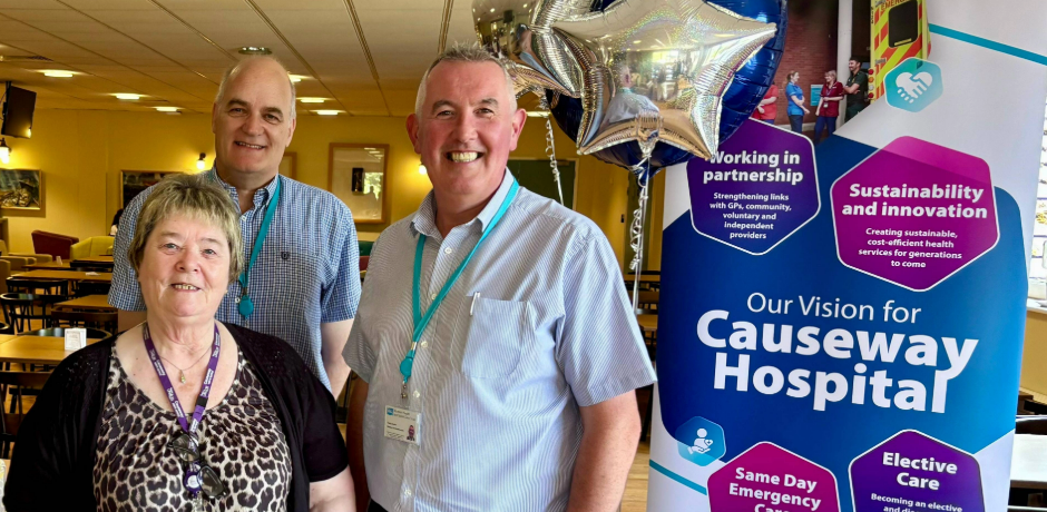 Three people stand smiling in front of a banner detailing the vision for Causeway Hospital, with blue and silver balloons.