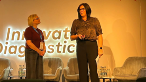 Two people, standing on stage, one is looking outwards, while the other stands to the side looking across.