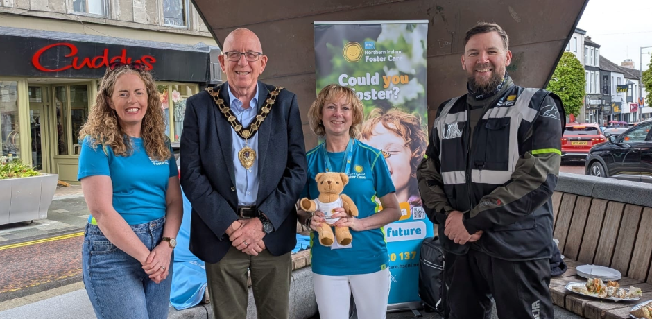 Two women wearing blue fistering-branded t-shirts, standing with a Mayor wearing a chain, and a biker, at the band stand in Ballymena.