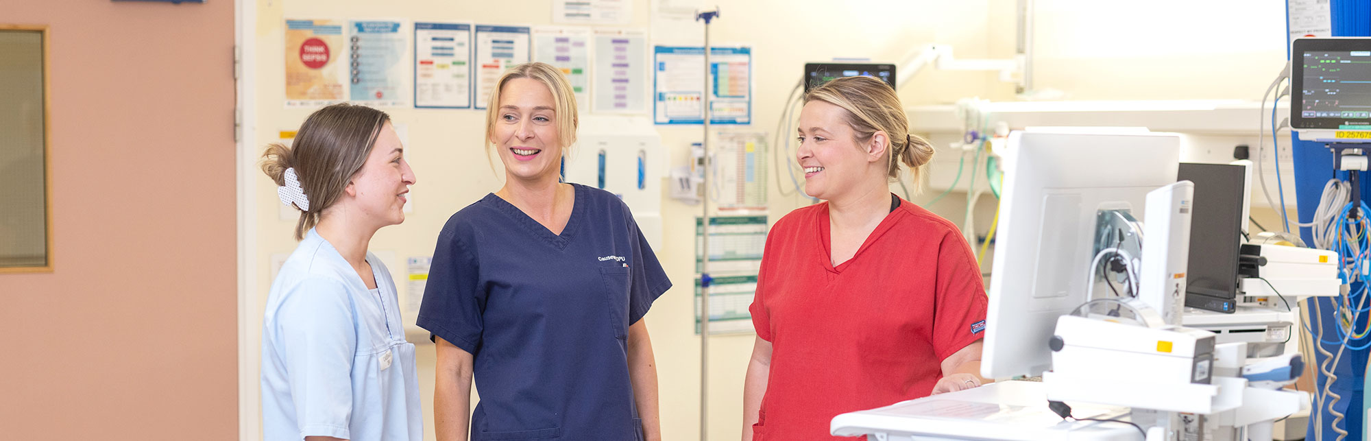 Three staff chatting in a hospital setting, one in light blue uniform, one in dark navy uniform and one in a red uniform.