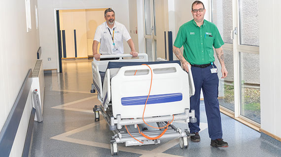 A health and social care staff member in white uniform with a porter in green t-shirt moving a bed along a corridor.