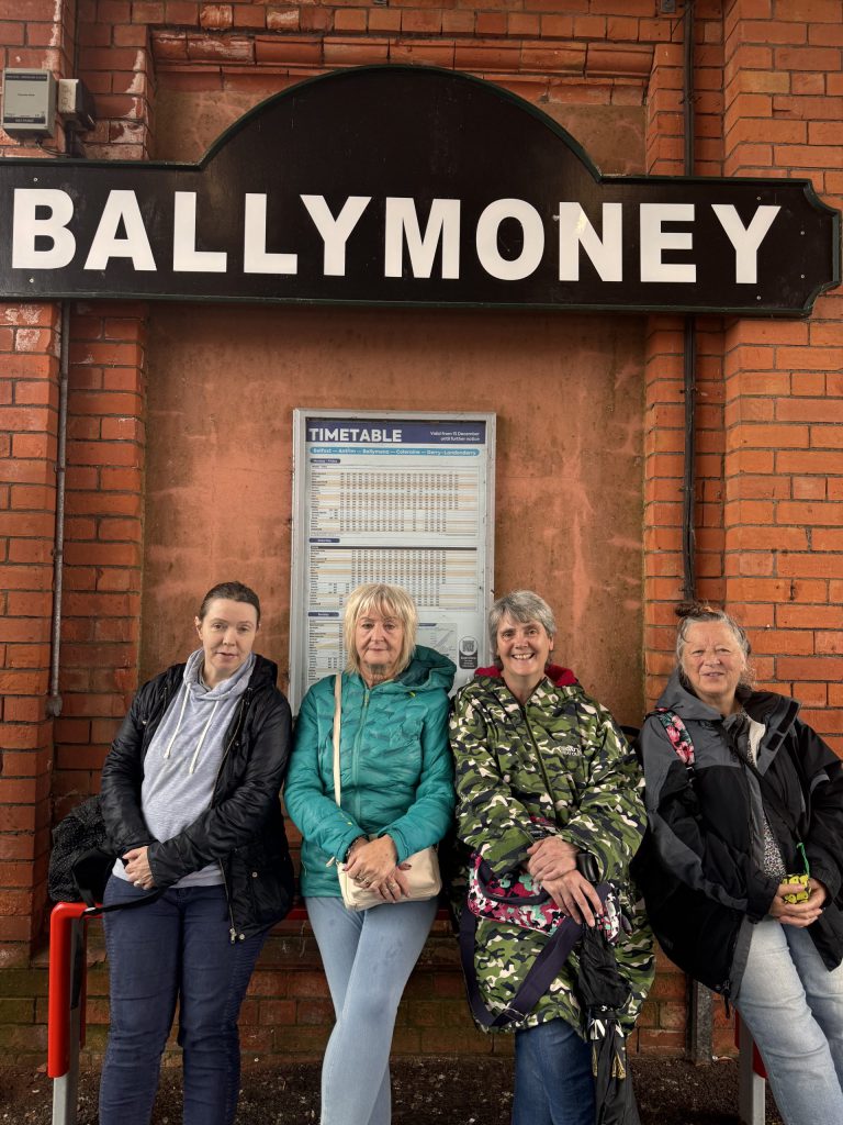 Four women sit on a bench with a large sign which reads 'Ballymoney' and a train timetable behind them.