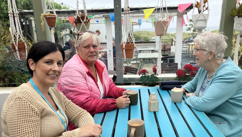 Three women sit at a blue table, with cups in front of them, in a cafe setting.