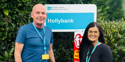 A man and a woman standing in front of an outdoor sign for Hollybank.
