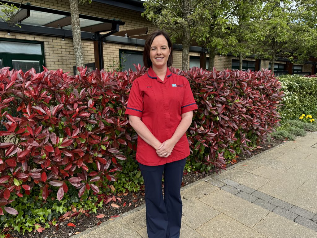 A nurse in a red tunic stands outside with a hedge and hospital building behind her.