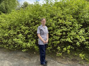 A nurse in a grey tunic stands outside with a hedge behind her.