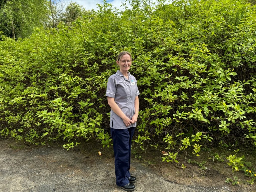 A nurse in a grey tunic stands outside with a hedge behind her.