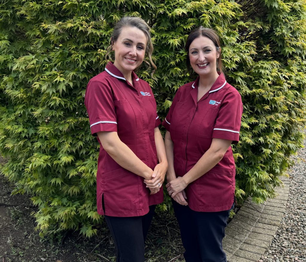 Two nurses in maroon tunic stands outside with a hedge behind them.