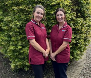 Two nurses in maroon tunic stands outside with a hedge behind them.