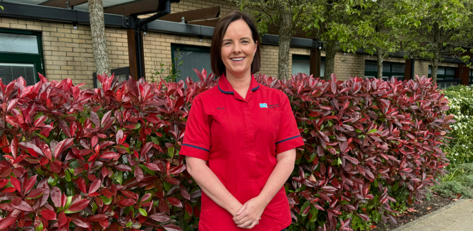 A nurse wearing a red tunic stands outside, with a hedge and hospital building behind her.