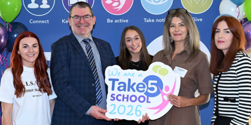 Five people standing together, holding a Take 5 sign, with balloons and Take 5 branding in the background.