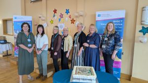 Seven women stand behind a table which has a large cake on it, with pop-up banners behind them.