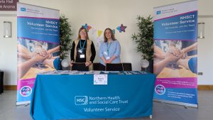 Two women stand behind a promotional table with pop-up banners either side of them.