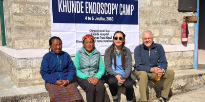 Two Nepalese women, Dr Amrita Gurung and a man sitting on a wall, with the sign behind them saying Khunde Endoscopy Camp.