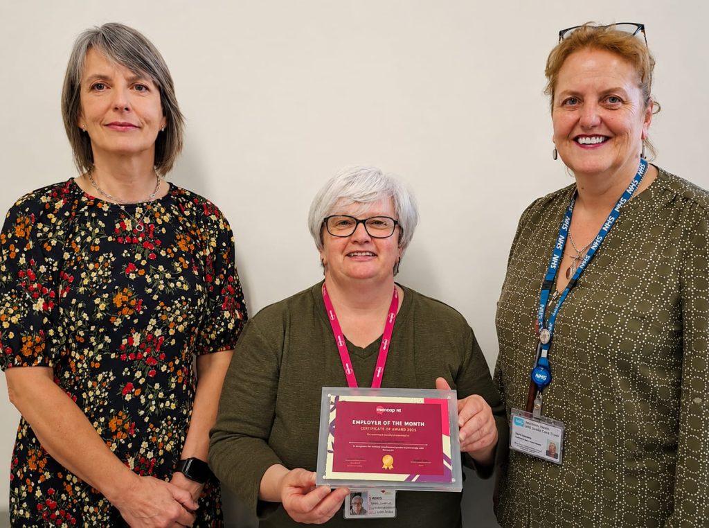 Three women standing together, smiling. The women in the middle is holding a glass 'employer of the month' plaque.
