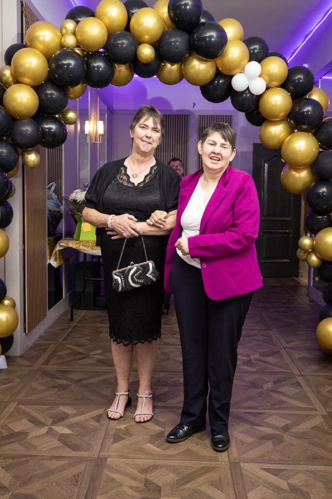 Two people stand in front of a balloon arch dressed for a formal occasion.