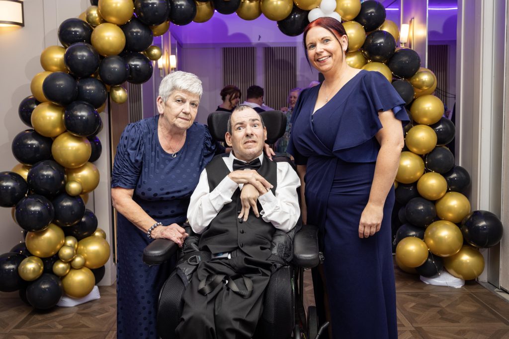 Three people, including one in a wheelchair, in front of a balloon arch dressed for a formal occasion.