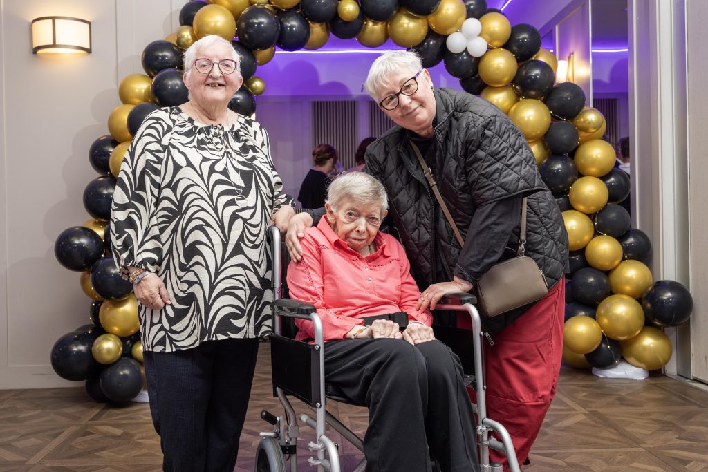 Three people including one in a wheelchair in front of a balloon arch dressed for a formal occasion.