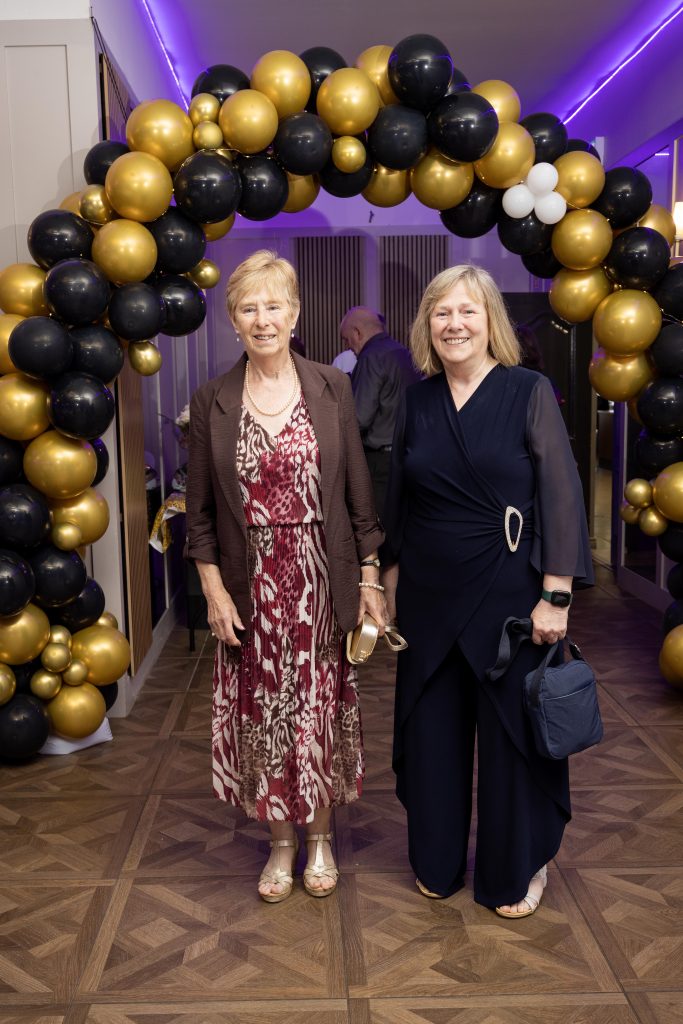 Two people stand in front of a balloon arch dressed for a formal occasion.