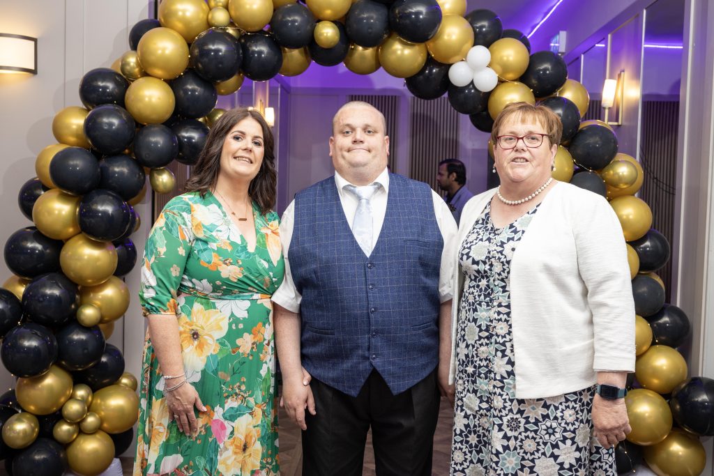 Three people stand in front of a balloon arch dressed for a formal occasion.