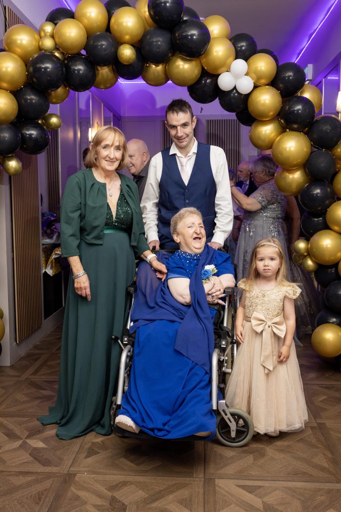 A group of people, including one lady in a wheelchair, stand in front of a balloon arch dressed for a formal occasion.