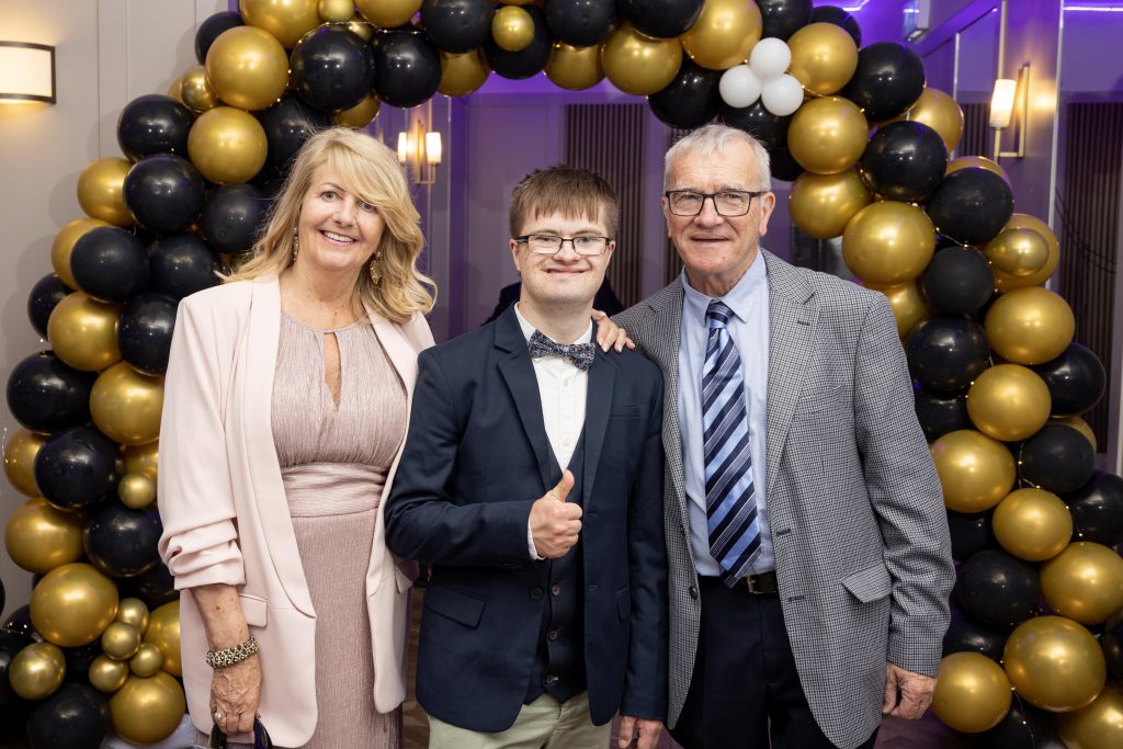 Three people stand in front of a balloon arch dressed for a formal occasion.