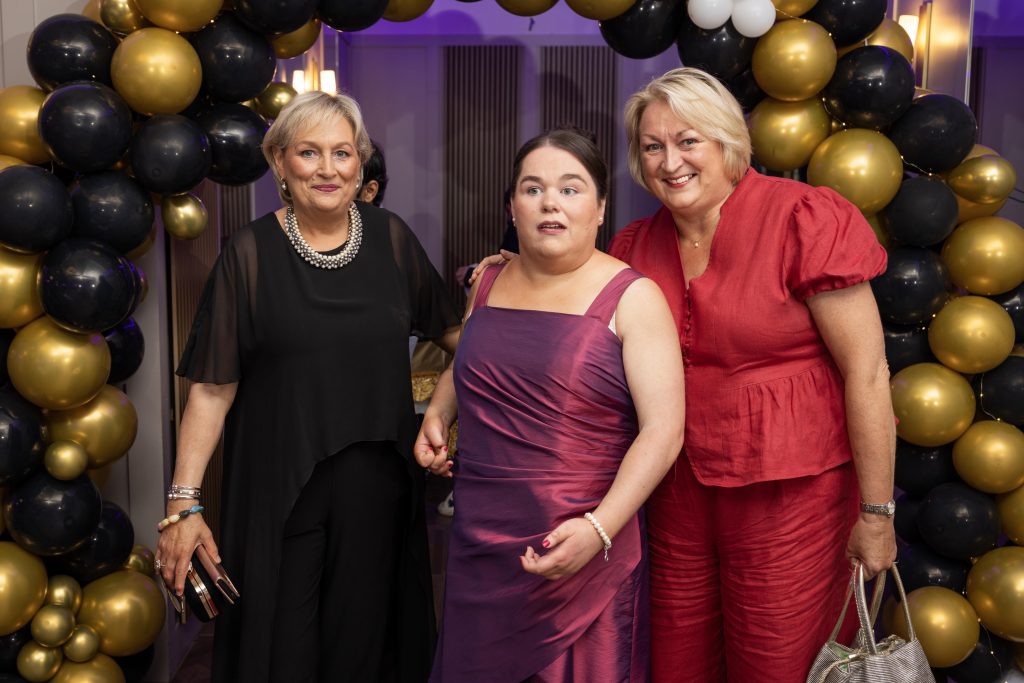 Three people stand in front of a balloon arch, dressed for a formal occasion.